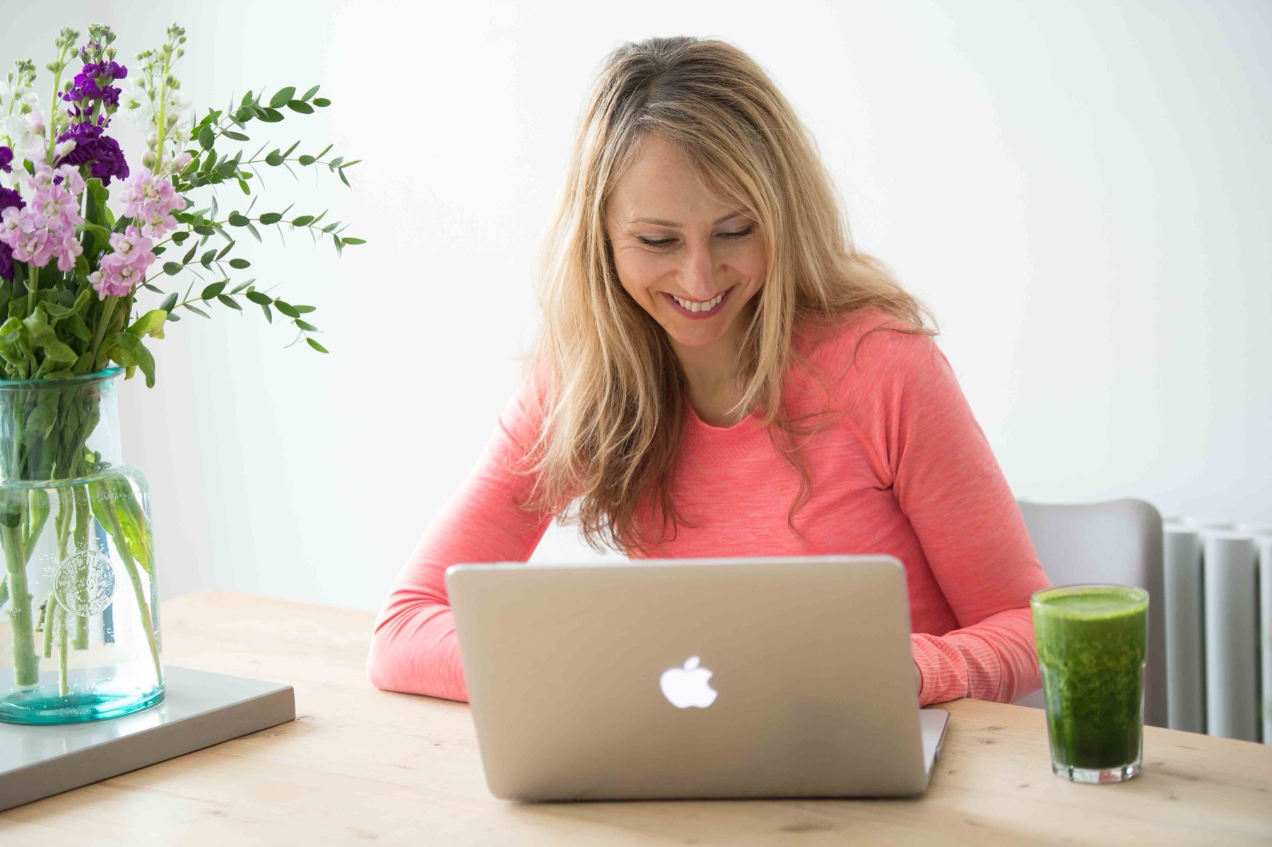Katie at a wooden table with her laptop, a jug of flowers and a green smoothie